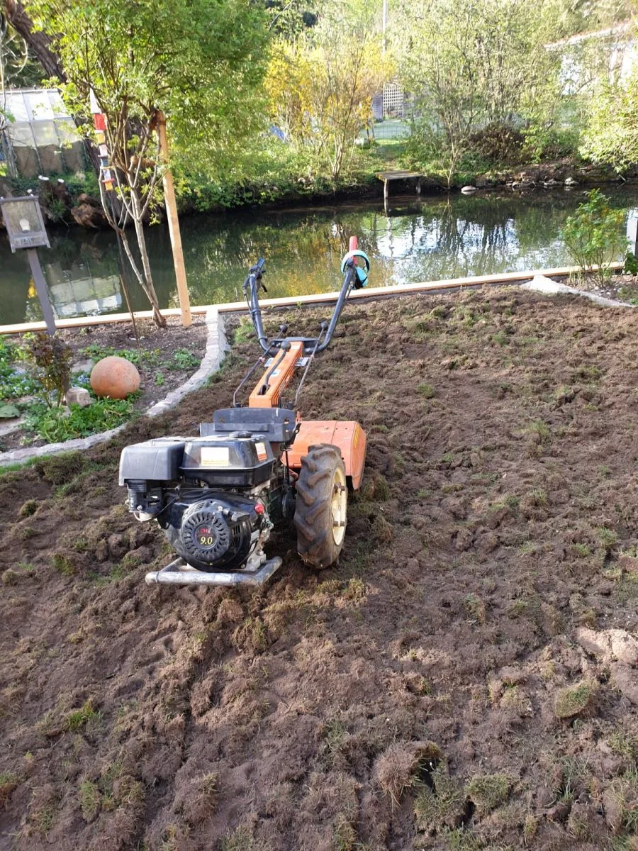 Motorisierte Bodenfräse beim Planieren einer Gartenfläche – orangefarbene Fräse auf frisch bearbeitetem Erdreich, Teich und Bäume im Hintergrund