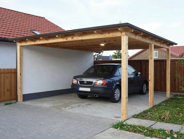 Holzcarport mit Pultdach und dunkler Trapezdacheindeckung vor Wohnhaus – Fahrzeug unter dem Dach auf gepflasterter Einfahrt