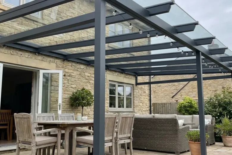 Terrassenüberdachung mit vollflächigem Glasdach und anthrazitgrauen Alu-Profilen als Hausanbau – Esstisch und Loungebereich darunter auf Natursteinbelag