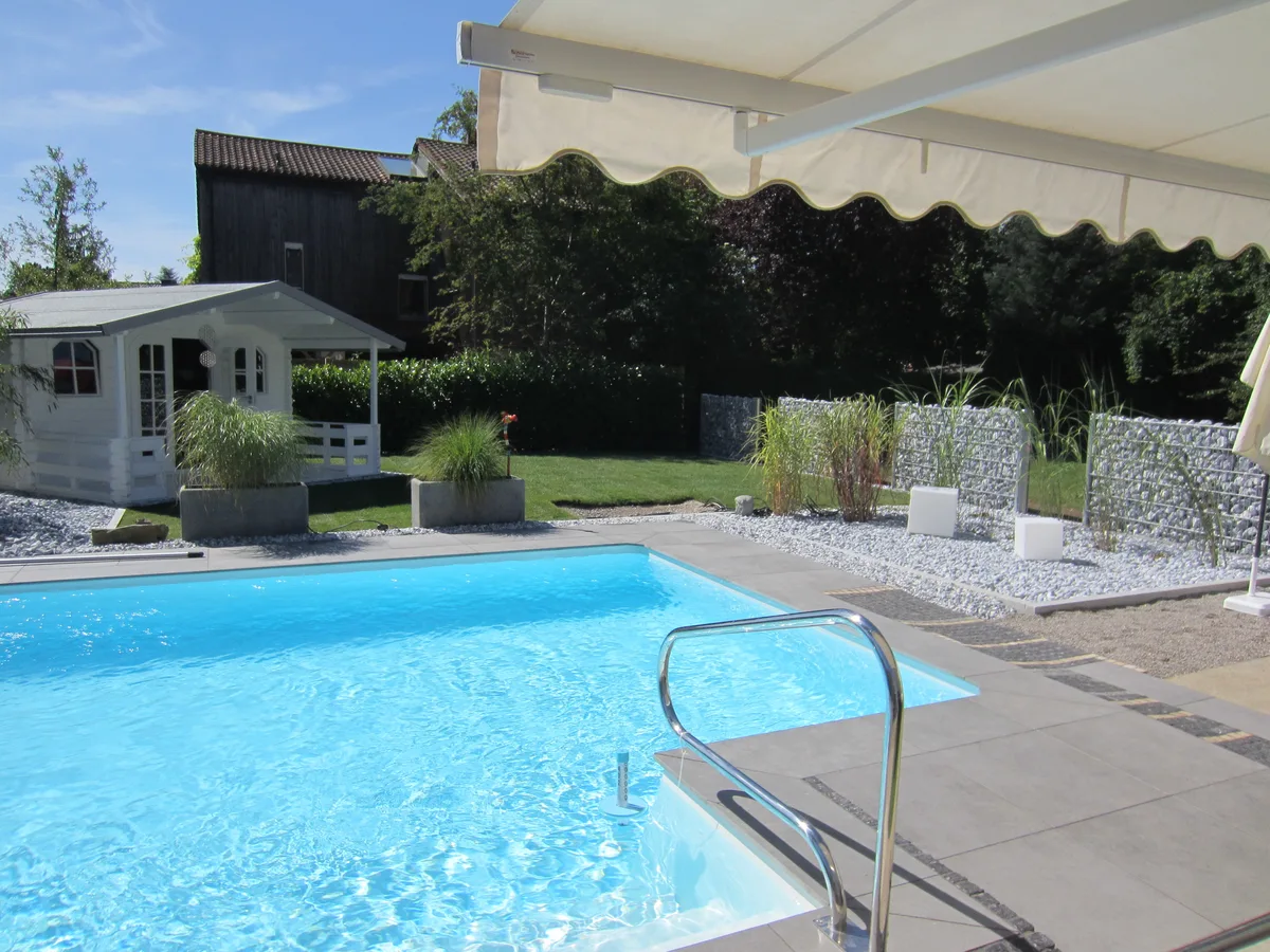 Fertiger Einbaupool im deutschen Privatgarten mit Edelstahlleiter, Gartenhaus, Kiesbeet und Markise – Blick vom Terrassenbereich