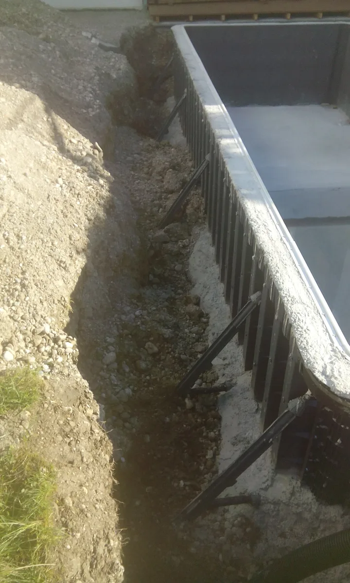 GFK-Einbaupool im Aushub mit montierter Stahlschalung an der Beckenwand – Hinterfüllungsbereich zwischen Becken und Erdreich vor dem Betonieren, Kiesboden und Rasenkante sichtbar