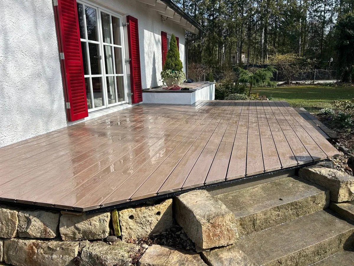 WPC Terrasse in Braun am Wohnhaus mit roten Fensterläden – nasse Dielen nach Regen, naturnahe Optik